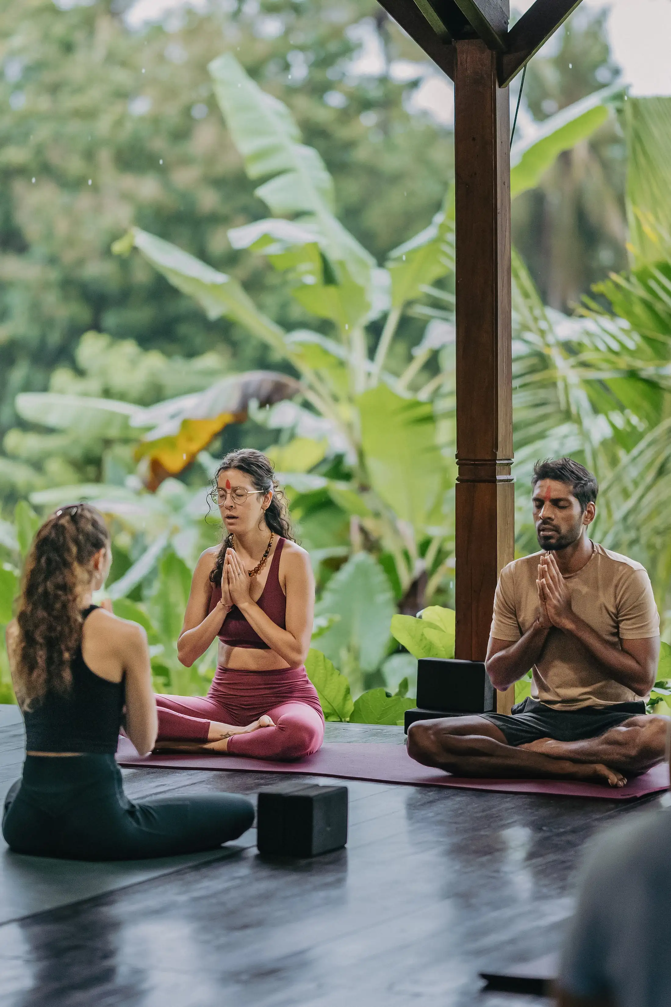 Two teachers and a student sitting cross-legged in prayer position on their mats in the open-air shala, lush jungle behind, at Senses in Ahangama