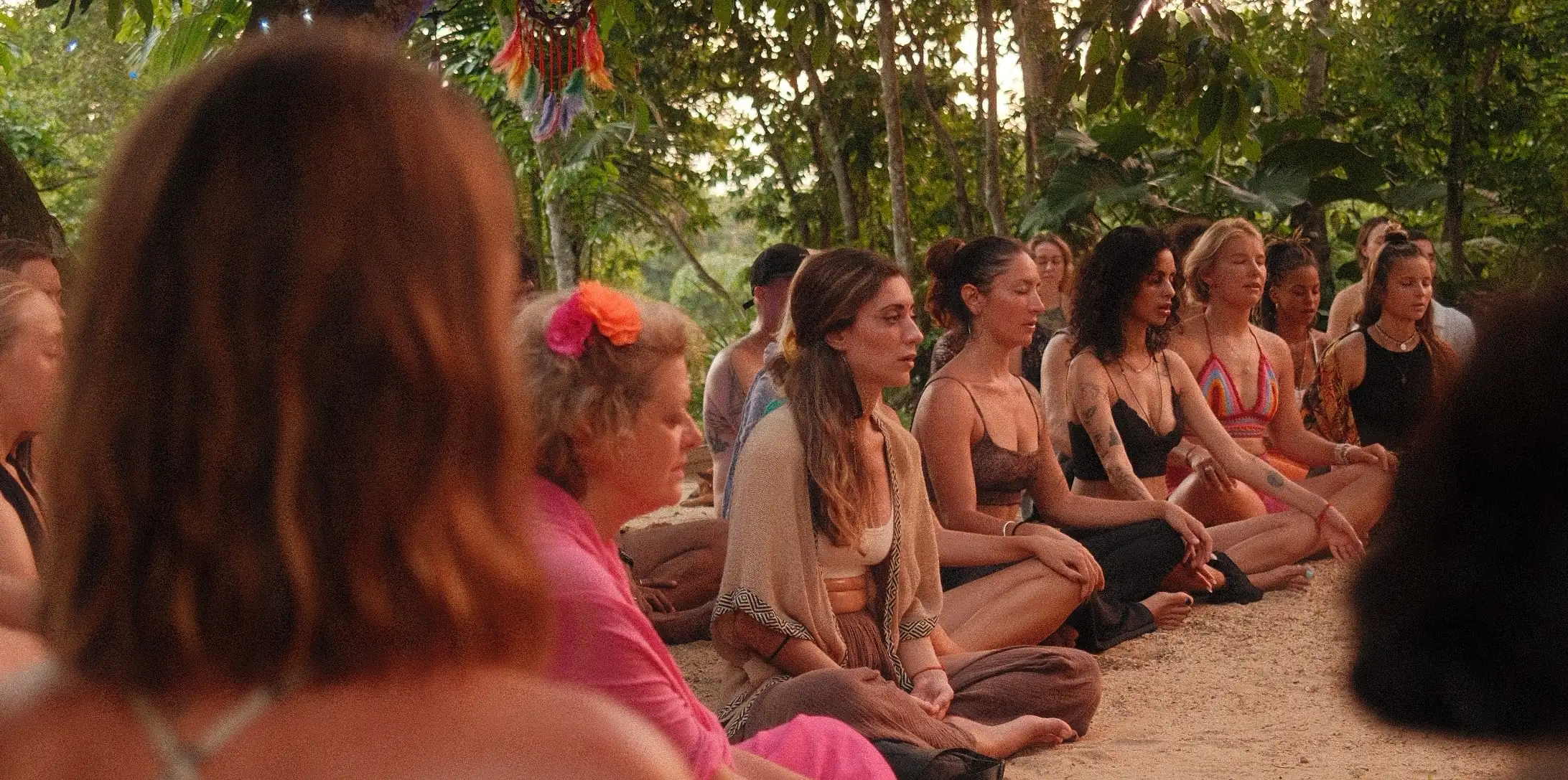 A row of people sitting cross-legged with their eyes closed on the sand under the trees at dusk, a feathered dreamcatcher hanging above them
