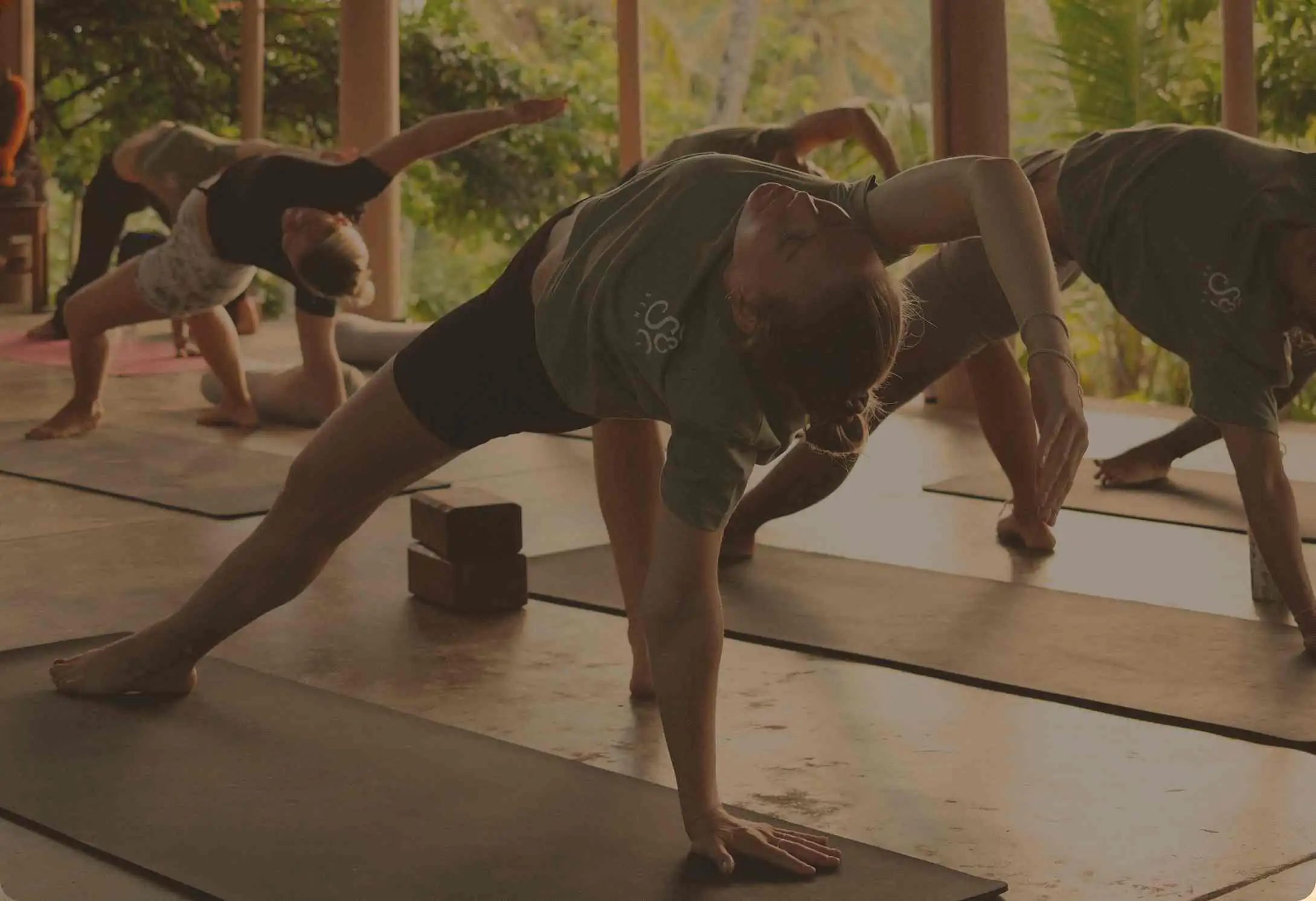 Trainees in olive Senses shirts moving through extended side angle pose on their mats in the open shala