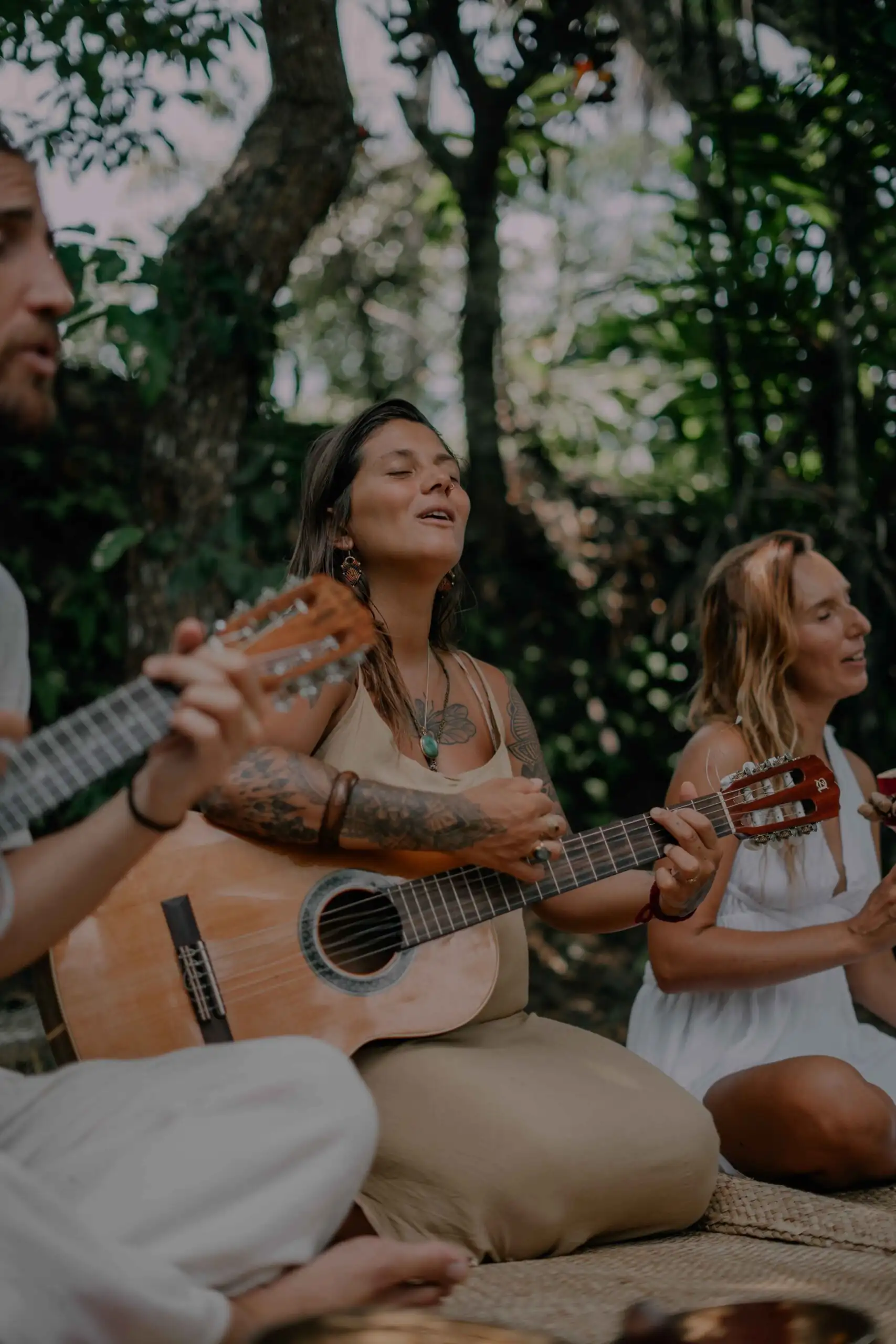 Three people sitting on woven mats under the trees at Senses, two of them playing guitar and singing with their eyes closed