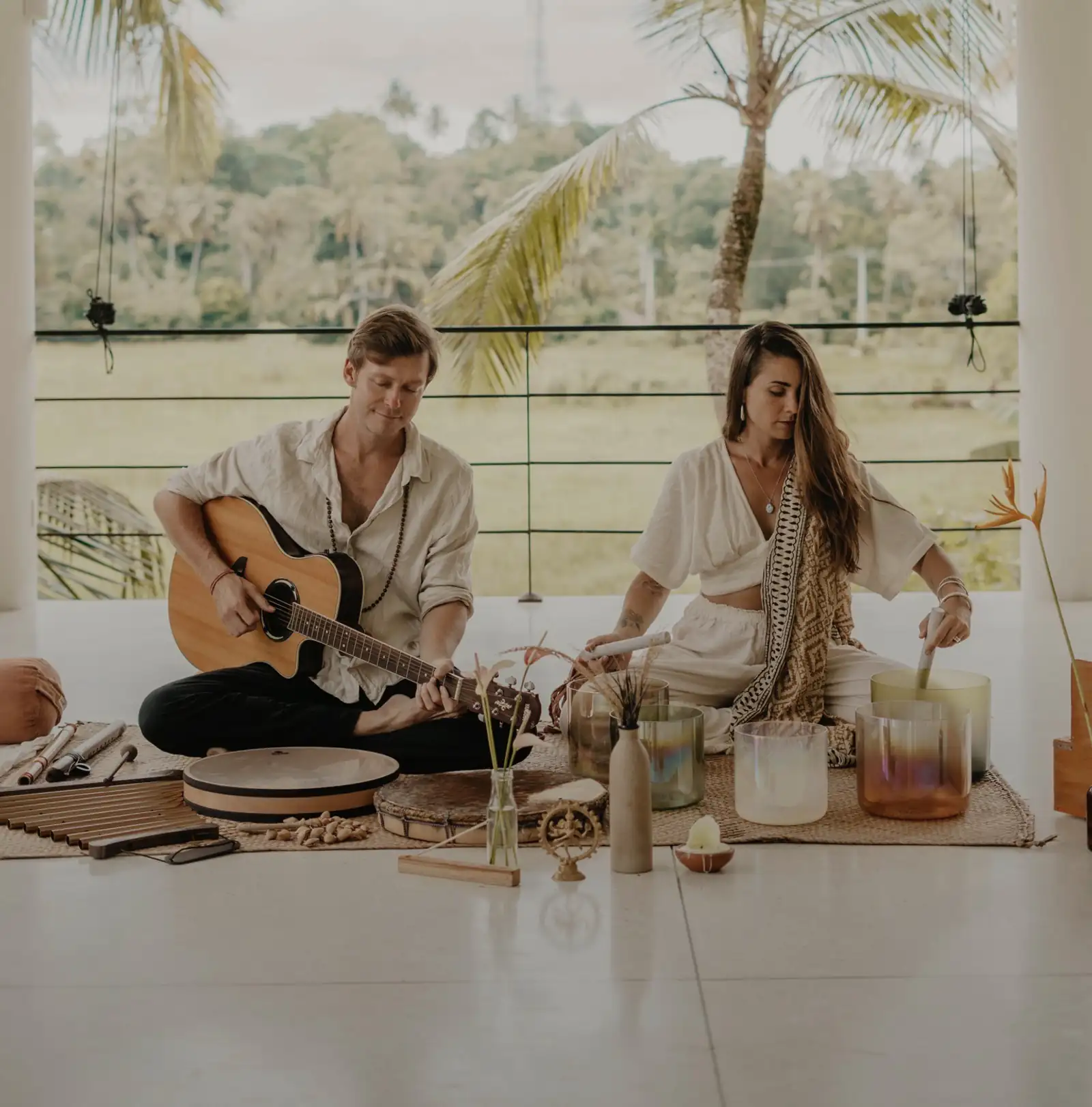A man playing guitar and a woman playing a crystal singing bowl on a white open terrace, with flutes, a frame drum and further bowls laid out around them and palm trees behind