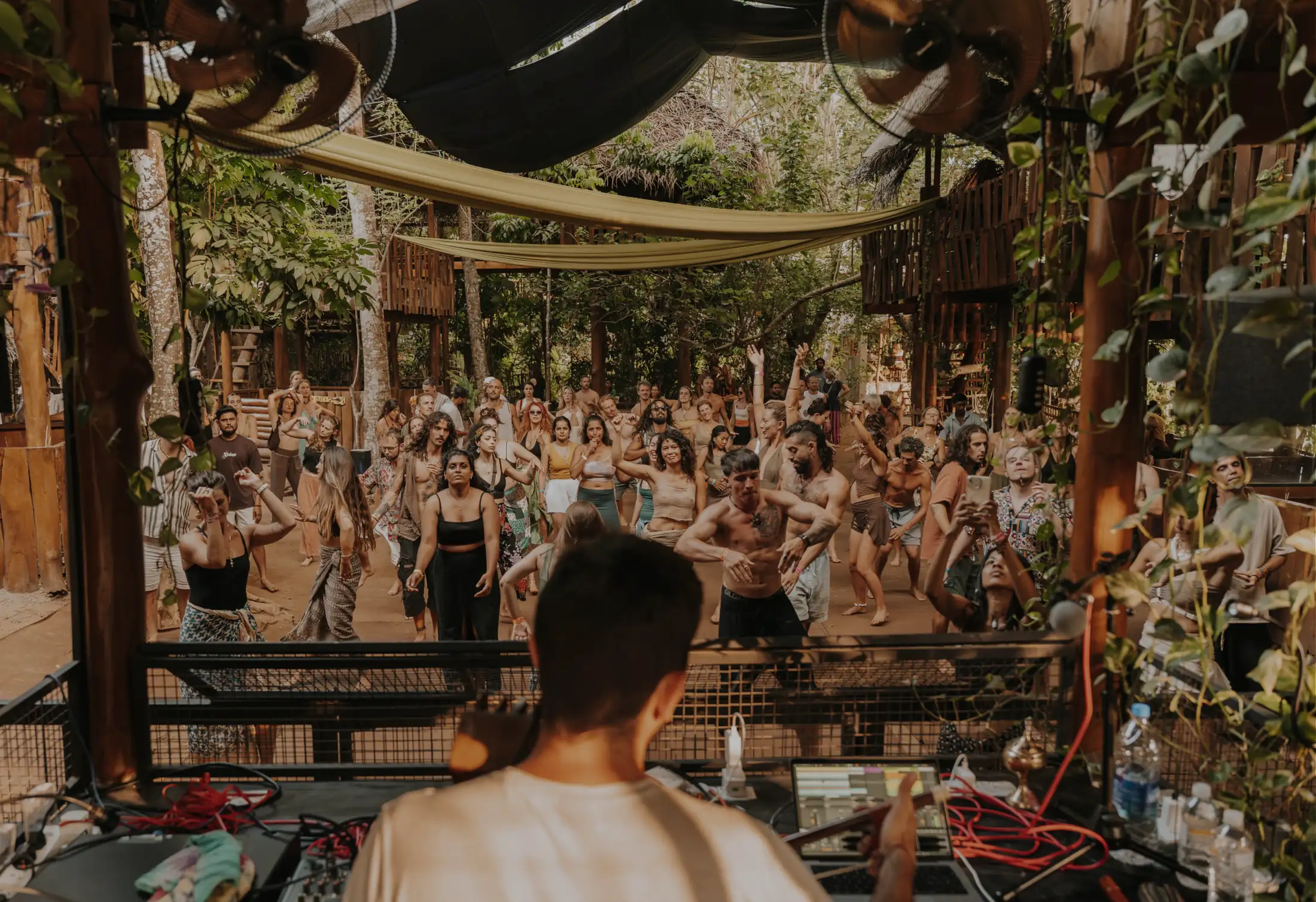 Seen from behind the DJ booth: a crowd dancing under fabric canopies in the open-air jungle venue in the afternoon light