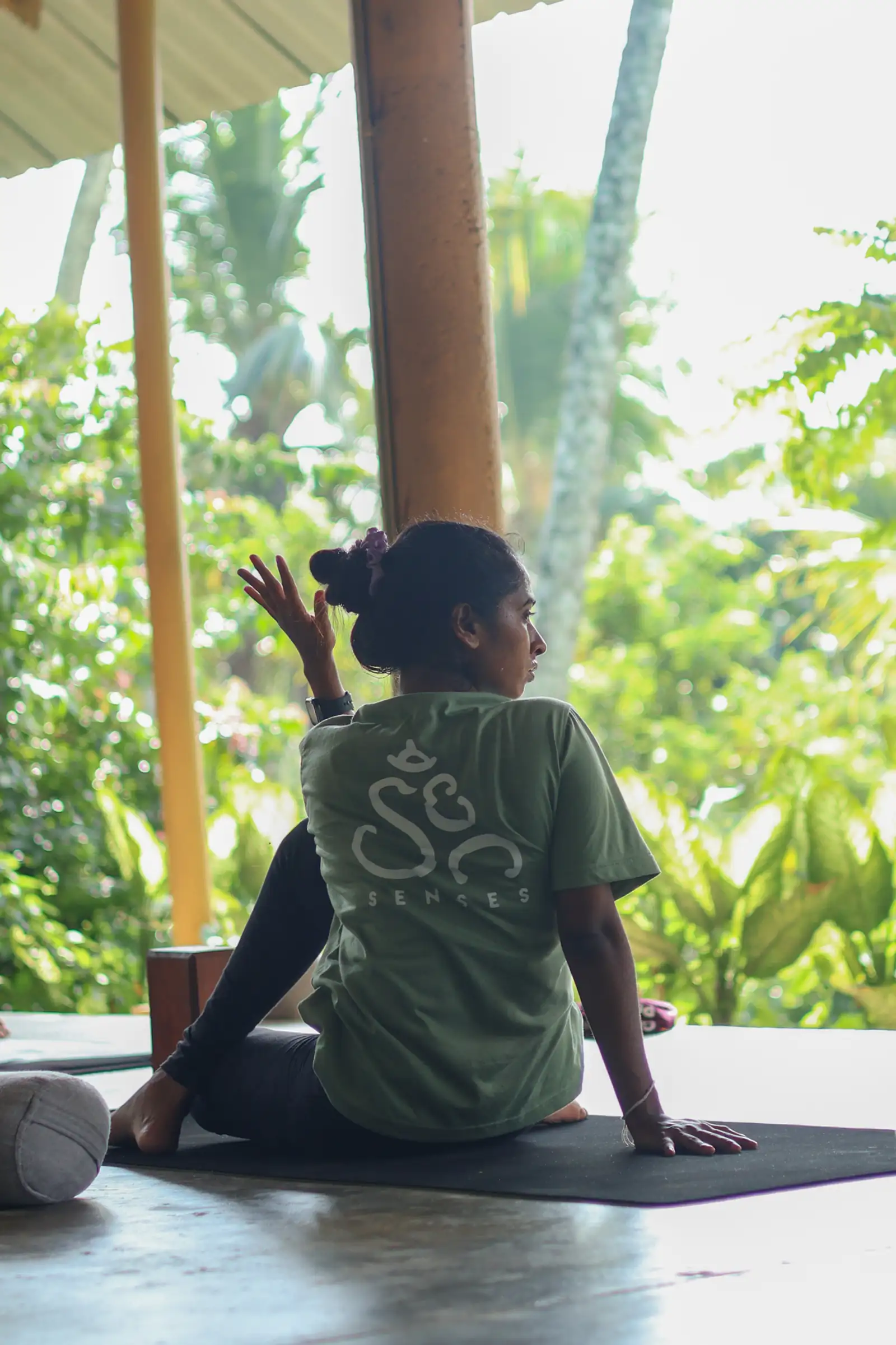 A Sri Lankan student in an olive Senses shirt practising a seated twist on her mat in the open shala, jungle greenery beyond