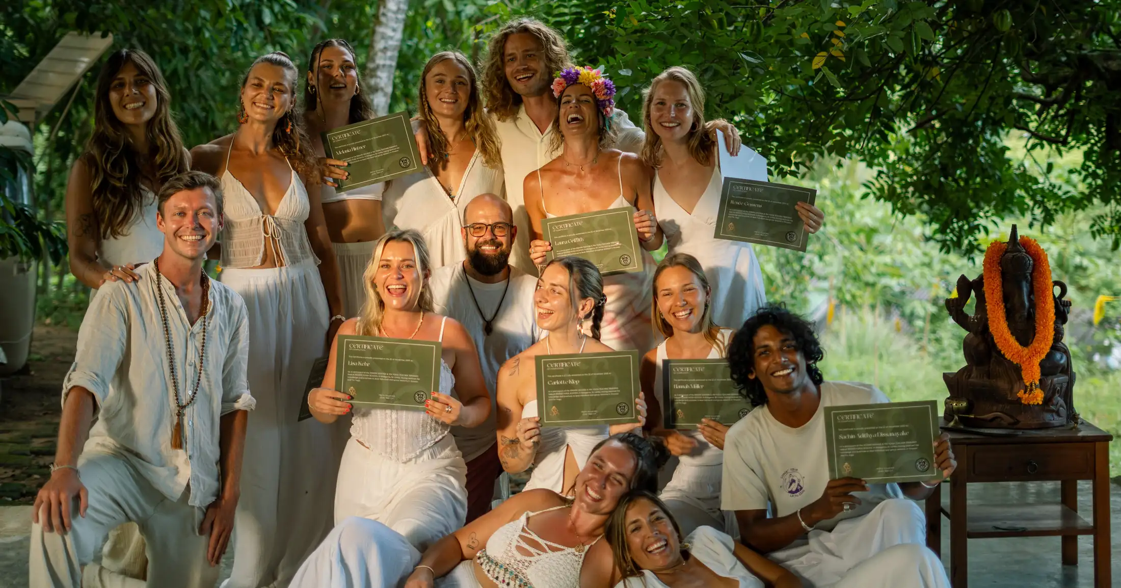 A graduating group in white holding their certificates and smiling together under the trees at Senses