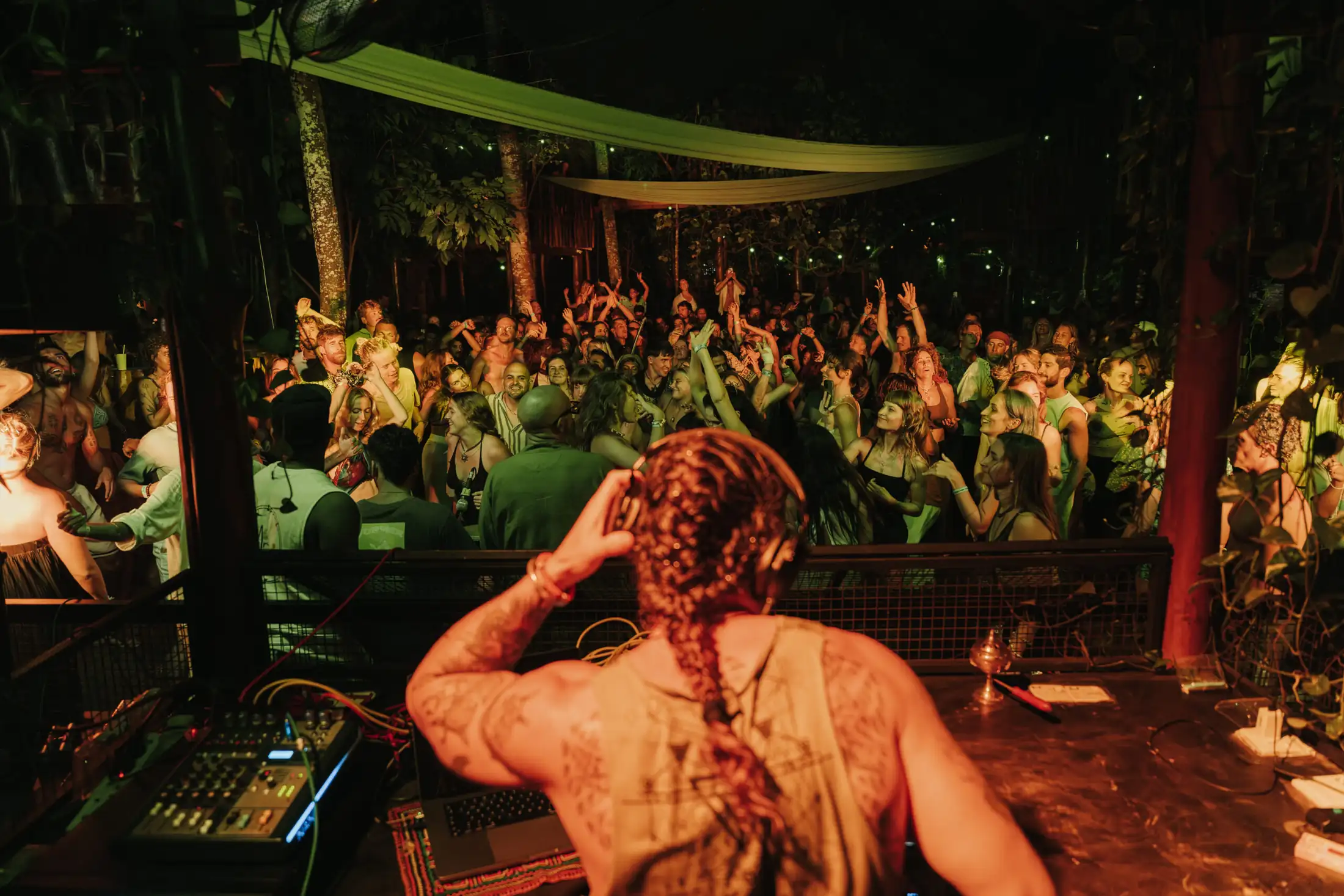 Seen from behind the DJ booth at night: a packed crowd dancing under fabric canopies strung between the trees, lit in green and orange