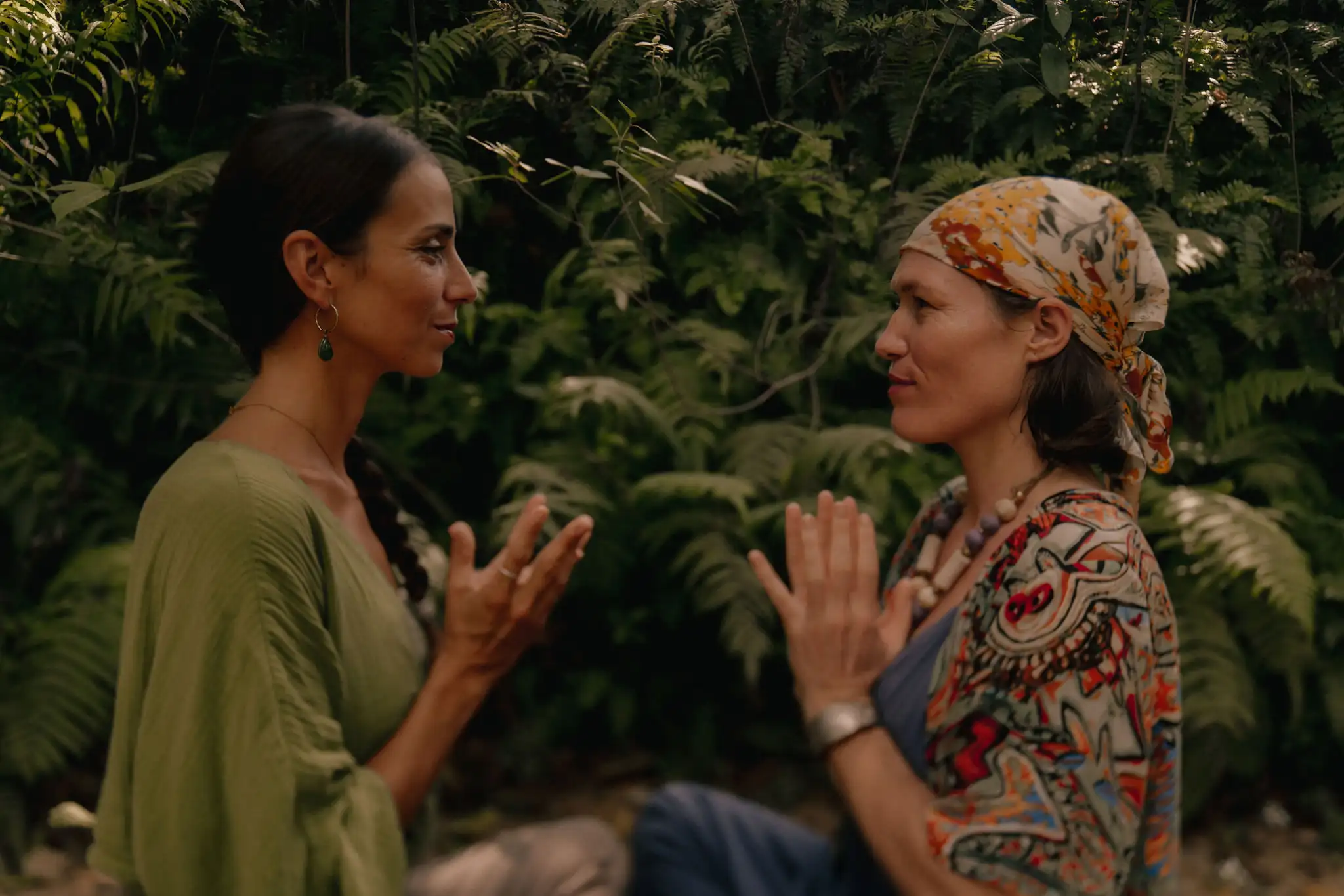 Two women sitting face to face in front of dense ferns, hands raised palm to palm in conversation